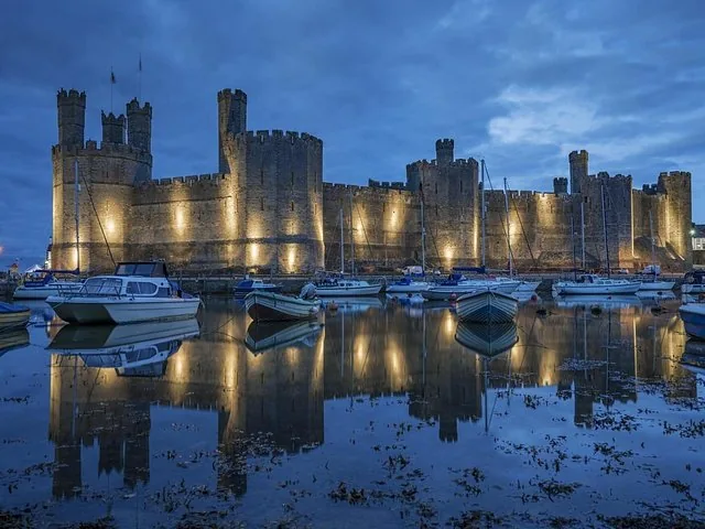 Il Castello di Caernarfon illuminato di notte che si riflette sull'acqua calma con piccole barche