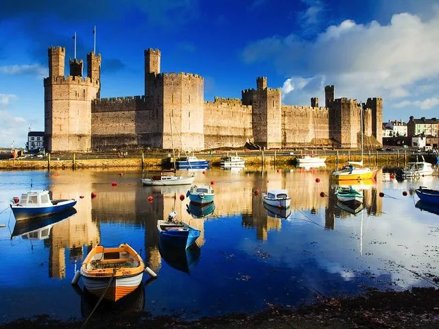 Caernarfon Castle an der Uferpromenade mit bunten kleinen Booten, die sich im Wasser spiegeln