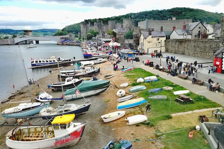 Barche adagiate sulla riva vicino al Castello di Caernarfon con turisti che camminano sul lungomare